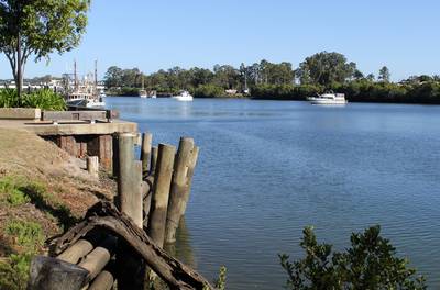 Peaceful view of the Burnett River with water, boats, wooden posts and trees under a clear blue sky in Bundaberg, Queensland, Australia